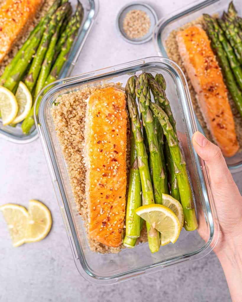 Woman's hand holding a glass container filled with salmon, asparagus, and quinoa.