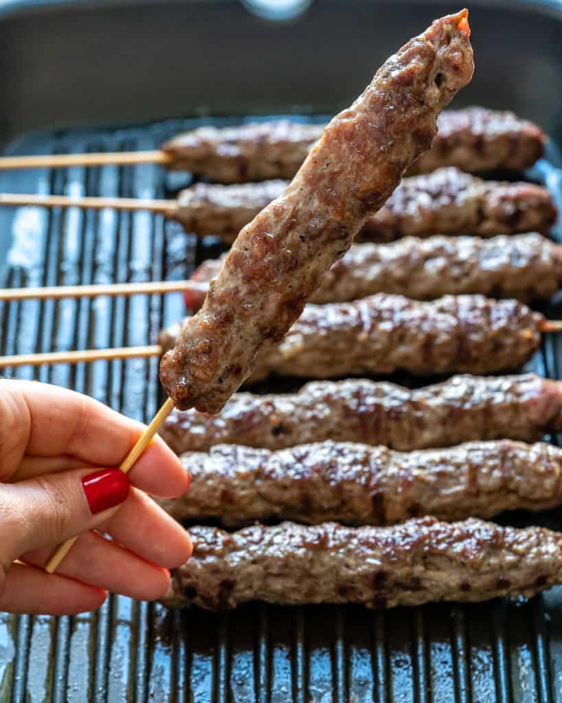 hand holding a grilled beef kafta skewer over a grill pan with other grilled kafta kabob