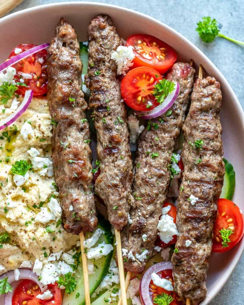 close up view of kafta skewers on a round plate with crumbled feta and slices fresh tomatoes with a side of hummus 