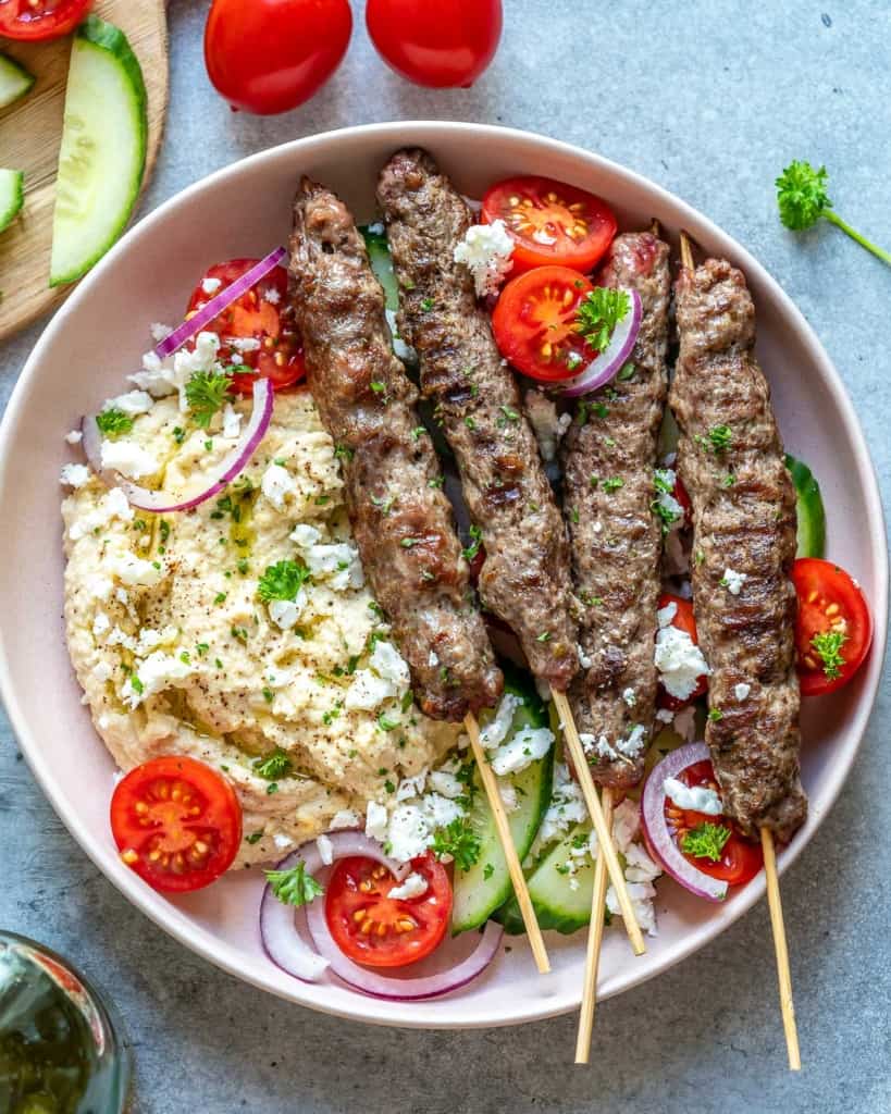 top view of 4 skewers of beef kafta on a round plate with salad and hummus