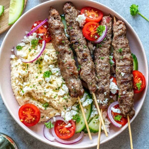 top view of grilled kafta skewers on a round plate with hummus, fresh tomatoes, and crumbled feta cheese