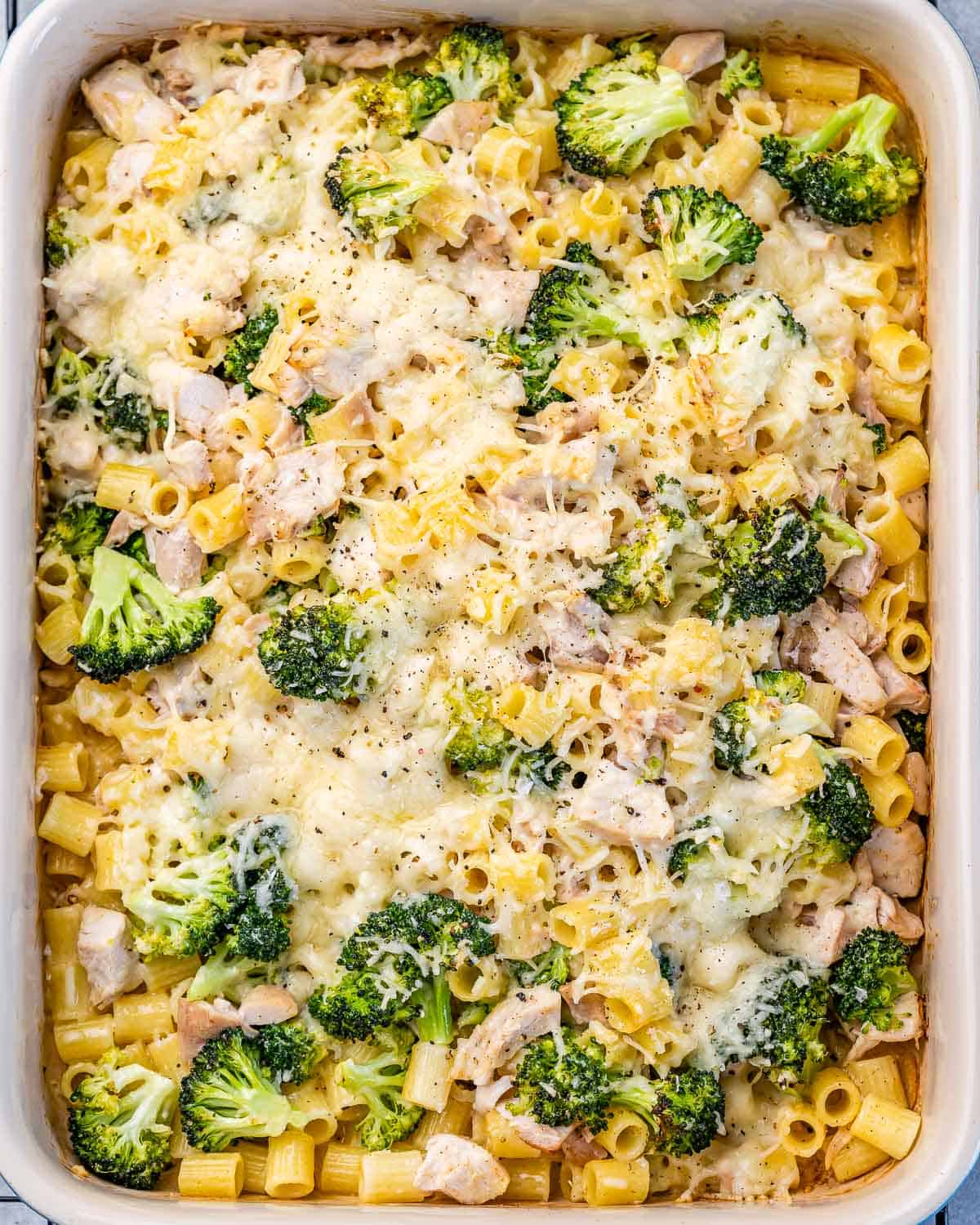 Chicken and pasta baked on a white dish with broccoli topped with cheese.