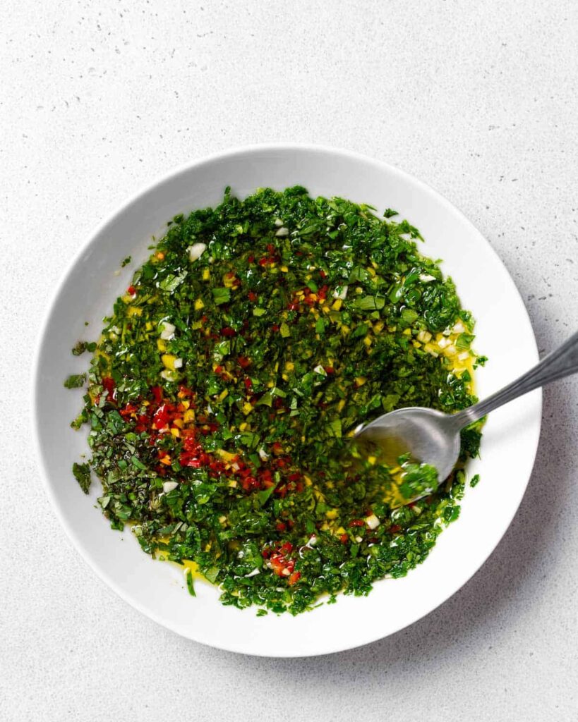 spoon mixing ingredients to make chimichurri wth oil in a bowl