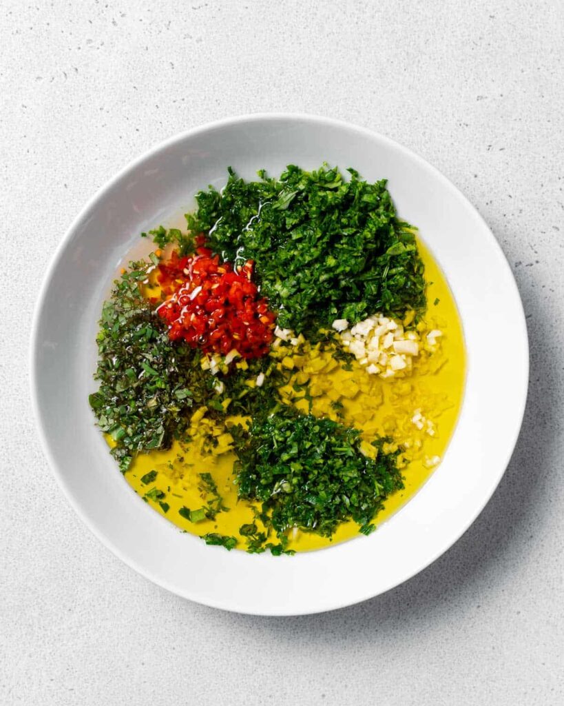 oil added to a white bowl with chopped cilantro parsley and peppers ro make chimichurri