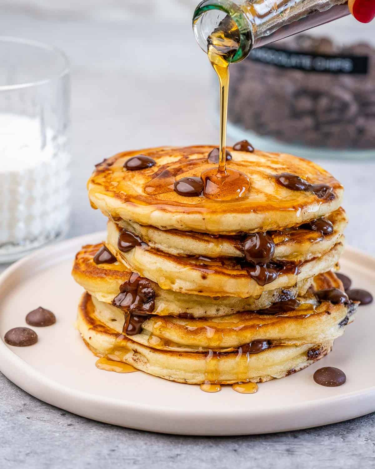 Drizzling maple syrup over a stack of chocolate chip pancakes on a white plate.