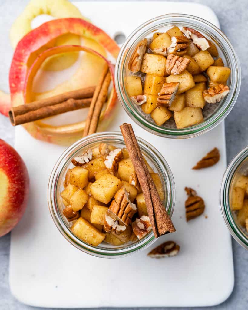 top view of chia puffin in two jars over a cutting board with cinnamon sticks and chopped apples on the side of the pudding jar
