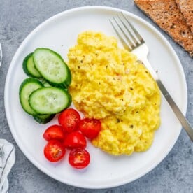 scrambled cottage cheese eggs with chopped cherry tomatoes and sliced cucumber on the left of the scrambled eggs.