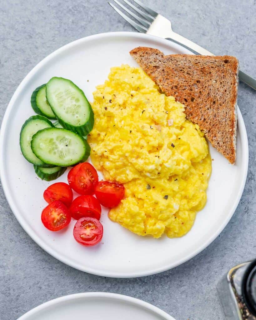 top view of a plate with scrambled eggs, sliced tomatoes, sliced cucumbers with a while grain toast bread by the eggs 