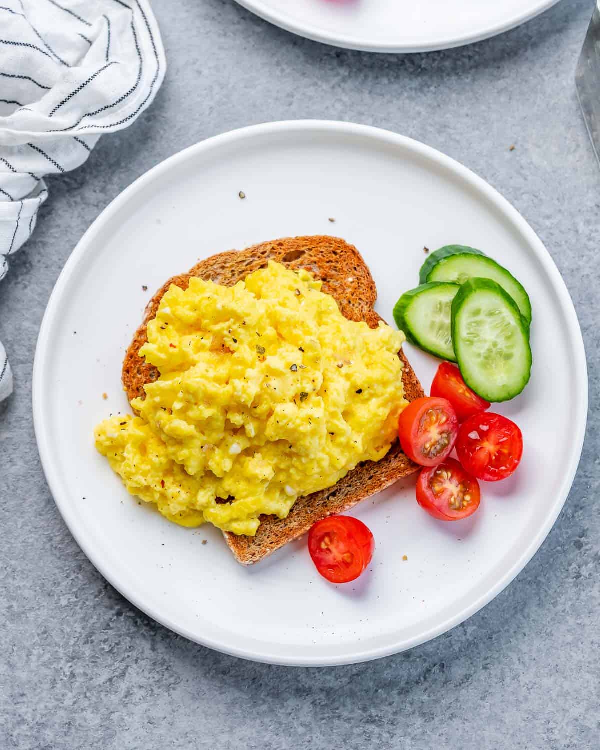 Scrambled eggs served on toast with a side of sliced cucumbers and grape tomatoes.