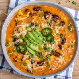 chicken enchilada soup in a round white bowl topped with shredded cheese and sliced avcoado.