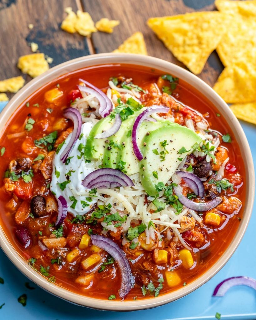 top view of a bowl of red chili with chicken beans and corn, topped with sliced avocado, sour cream, and shredded cheese