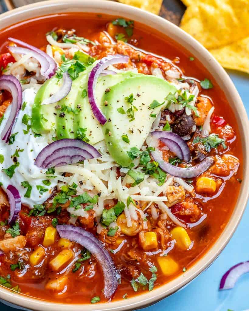 close up image os a bowl of chicken chili with black beans, corn, and chicken, topped with sliced onions, sliced avocado and shredded cheese