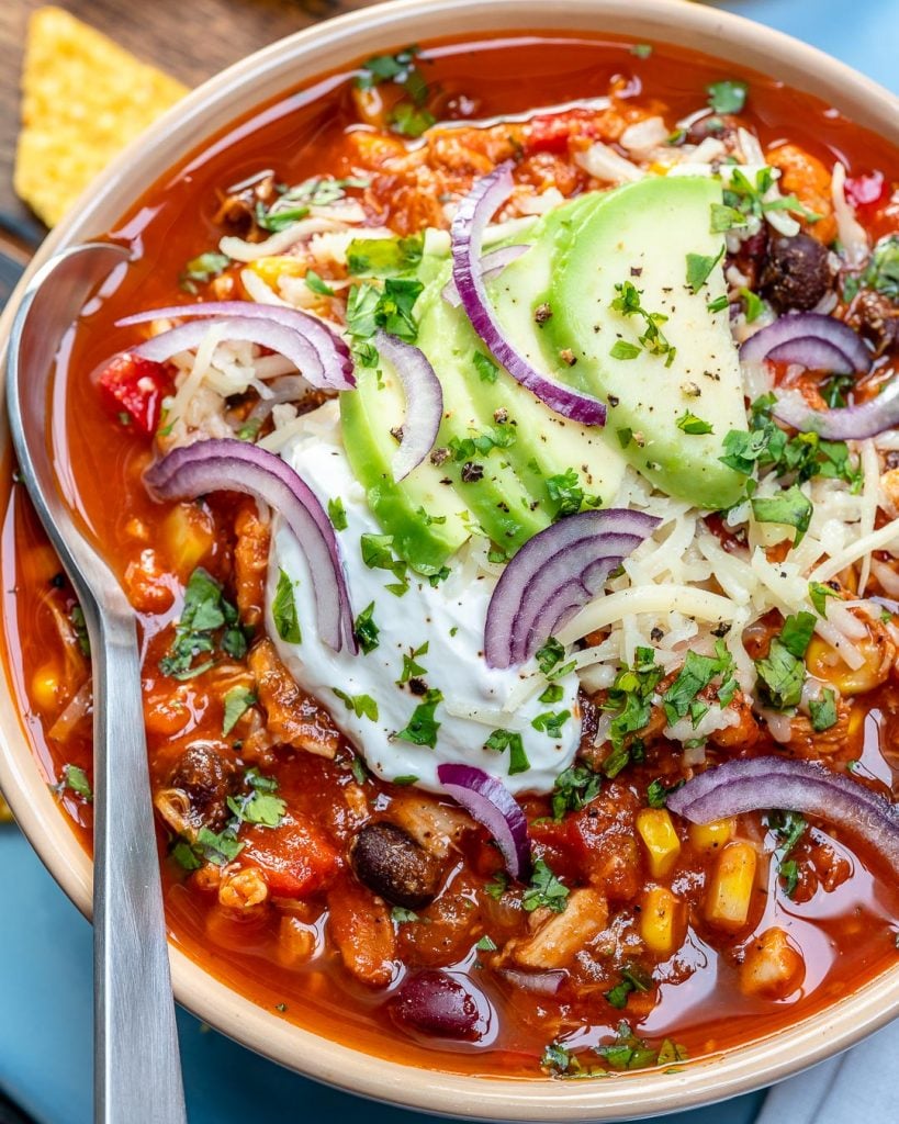 Top view of a white bowl with red looking chicken chili with spoon in bowl, topped with sliced avocado, sour cream, and shredded cheese