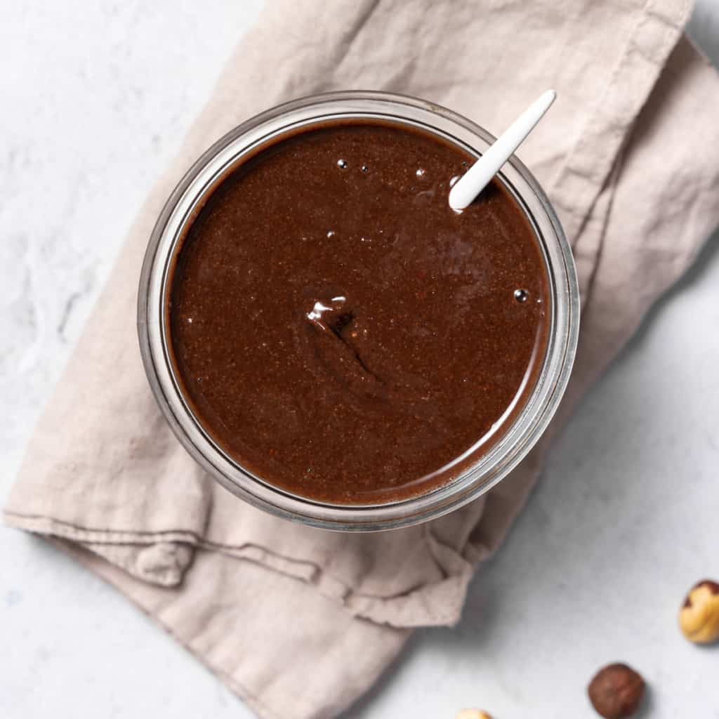 top view of a small bowl with brown colored homemade nutella with spoon in jar