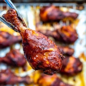 hand holding one chicken drumstick over pan with many baked drumsticks