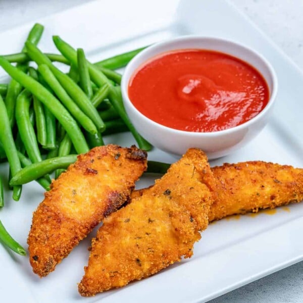 a plate with 3 chicken tenders and a side of green beans with a smal bowl of ketchup
