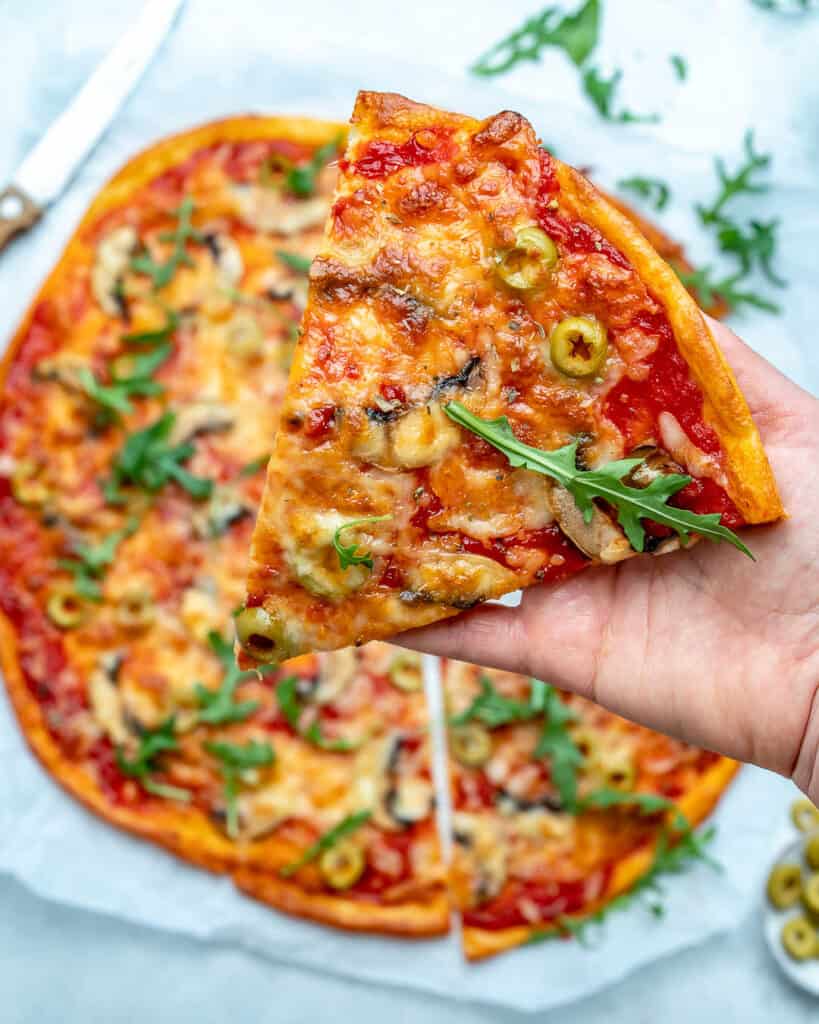 hand holding one slice of low carb pizza over a full pizza that it stopped with fresh arugula and green sliced olives 