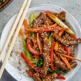 top view of a white bowl of white rice topped with Mongolian beef with chop sticks on the left of the bowl