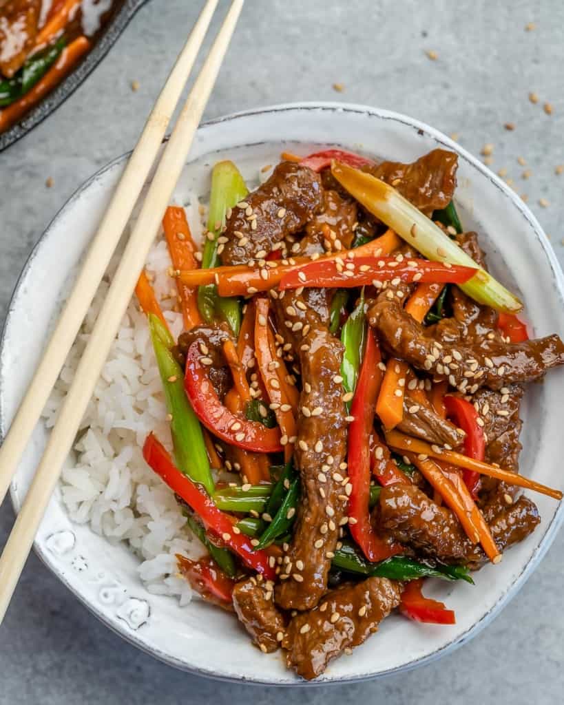 top view of a white bowl of white rice topped with Mongolian beef with chop sticks on the left of the bowl