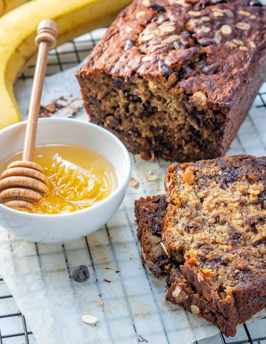 Oatmeal chocolate chip banana bread on a rack net to a bowl of honey with some of the banana bread sliced.