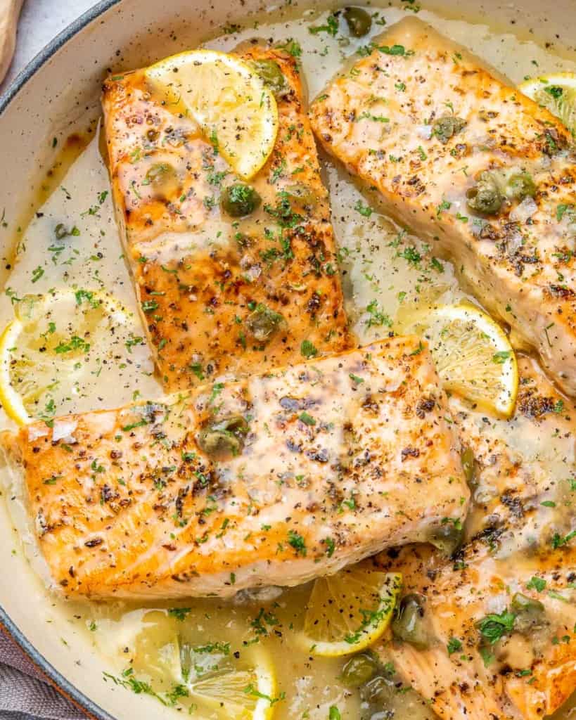close up side shot of seared salmon filets in a lemon sauce topped with capers and lemon wedges
