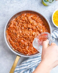 Pouring pasta water into a pan with ground beef and sauce.
