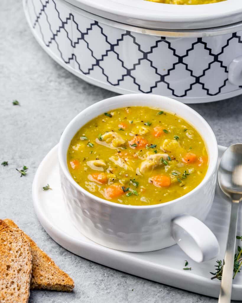 side shot of a white cup with split pea soup next to a crockpot