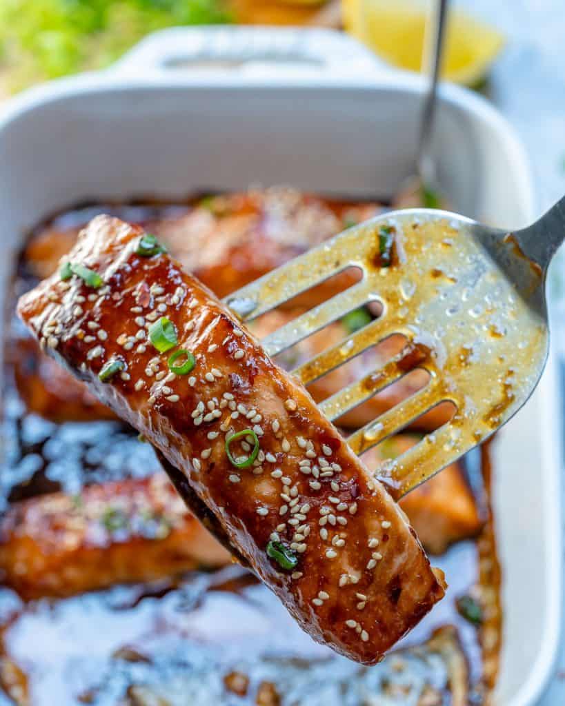 metal spatula holding a piece of baked teriyaki salmon that is garnished with sesame seeds and sliced green onions