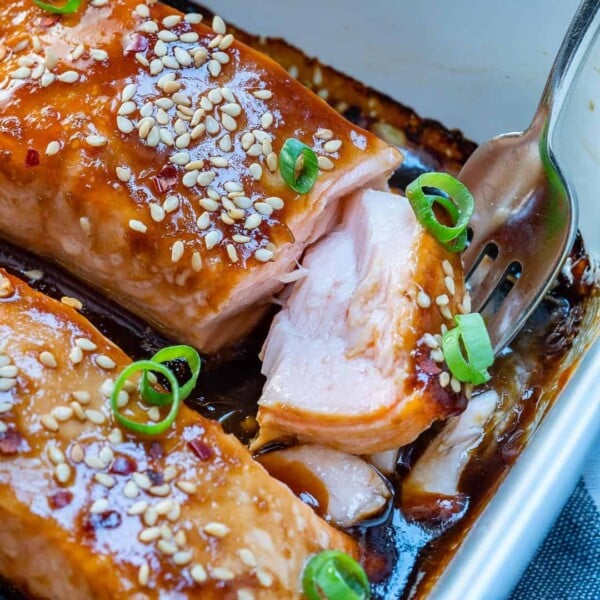 side shot of a fork getting a morsel of the baked teriyaki salmon from a white baking dish