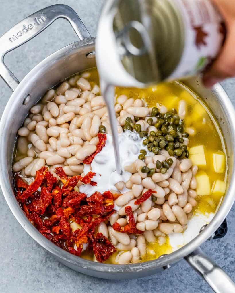 Pouring coconut milk into soup pot with white beans and potatoes.